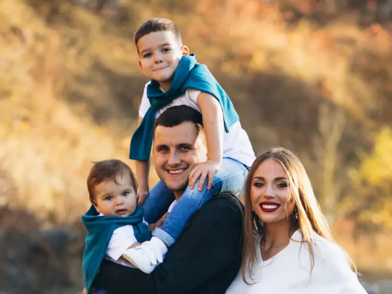 A family of four poses outdoors, with two children sitting on an adult's shoulders, dressed in casual attire amid a natural backdrop.