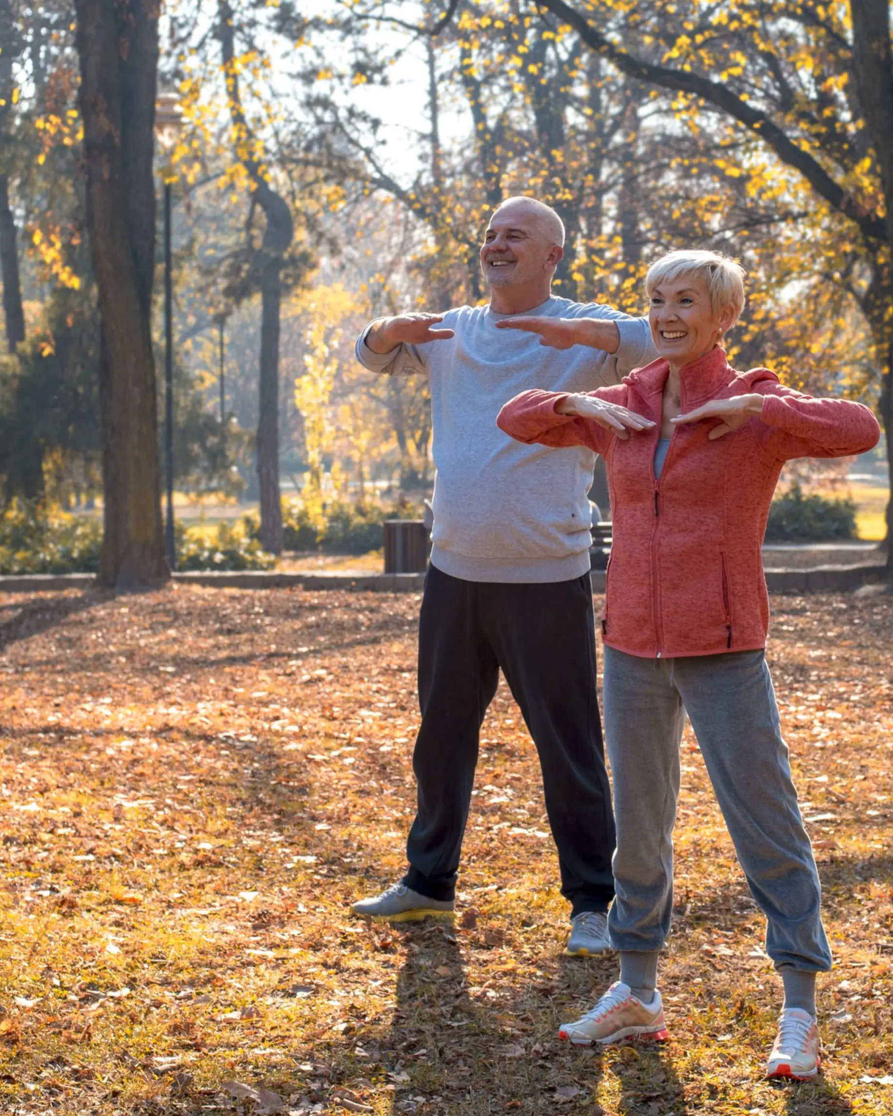 A man and a woman stand in a park during autumn, practicing stretches amid fallen leaves and trees with golden foliage.