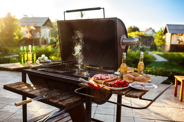 1836 A grill with smoky barbecue, surrounded by bottles of beer, burgers, sausages, and fresh veggies, set in a sunny backyard.