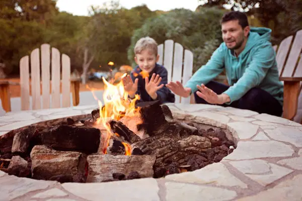 Firepit Stock Image A cozy campfire surrounded by stone, with a child and adult warming their hands, seated on wooden chairs in a serene outdoor setting.